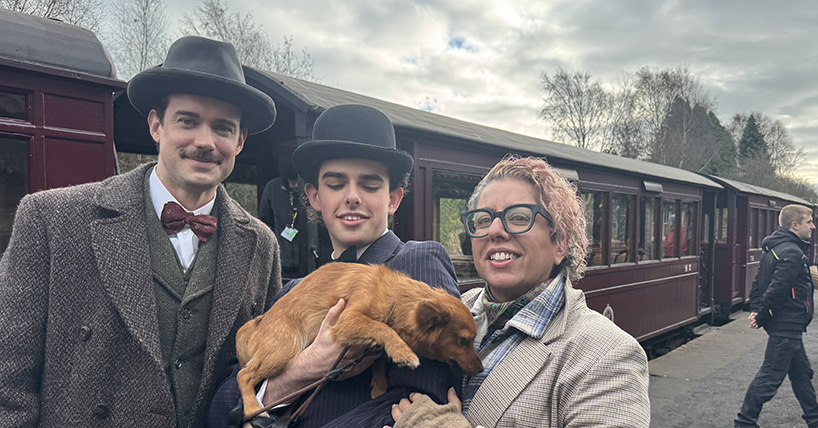 2 actors in period costume holding a brown dog alongside Tina Gharavi with a steam train in the background 2 actors in period costume holding a brown dog alongside Tina Gharavi with a steam train in the background